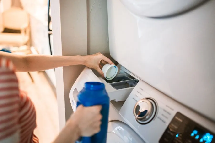 Person pouring laundry detergent into a front-loading washing machine connected to a laundry drain line